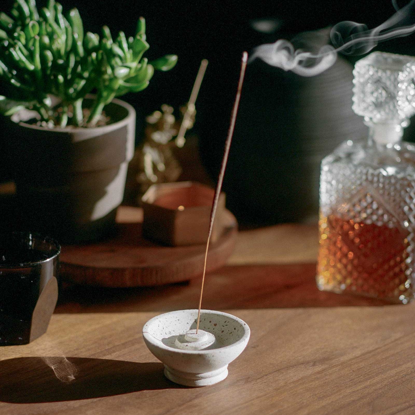 Incense stick burning in a small white dish on a wooden surface with plants and a glass container in the background.