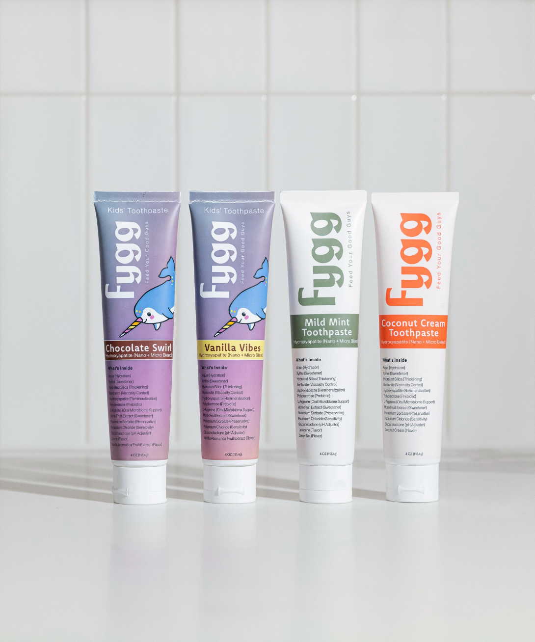 Four Fugg toothpaste tubes standing upright on a white surface against a white tiled background. The tubes are labeled Chocolate Sweets, Vanilla Vibe, Mint Mint, and Coconut Cream, with purple, lavender, white, and white-orange packaging.